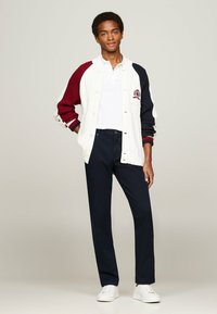 Giacca lavorata a maglia bianca e bordeaux con maniche blu navy, indossata sopra una polo bianca, abbinata a pantaloni blu navy e sneakers bianche.