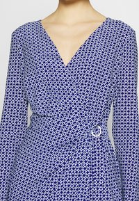 Abito blu avvolgente con un motivo geometrico bianco, scollatura a V profonda, maniche lunghe e una cintura in tessuto fissata con una fibbia metallica circolare.