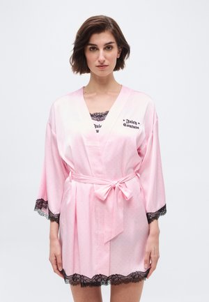 Vrouw draagt een roze satijnen badjas met zwart kanten randje, gestrikt in de taille, met de tekst "Juicy Couture" op de borst.