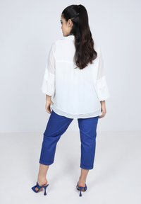 Weiße, durchscheinende Bluse mit geschichteten Rüschem-Ärmeln, kombiniert mit knöchelhohen, kobaltblauen Hosen und schwarzen, Absatzsandalen. Insgesamt glatte Textur.