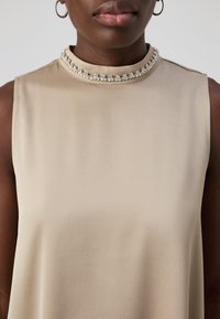 Vaaleanbeige ilmanhiha blouse, jossa on korkea kaulus, ja koristeellinen kristallireunus kauluksessa. Silkkinen, kevyt kangas.