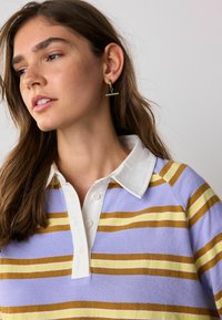 Donna con capelli castani fino alle spalle che indossa orecchini a croce dorati e una camicia a righe viola, gialla e bianca con colletto, che guarda a sinistra.