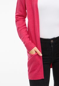 Long cardigan in pink, made of soft fabric. Features ribbed cuffs and a side pocket, paired with black high-waisted jeans.