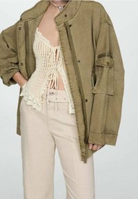 Olijf groene oversized jas met een ritssluiting, voorzien van een opvallende kraag en zijkanten met zakken, gelaagd over een crèmekleurige gehaakte top.