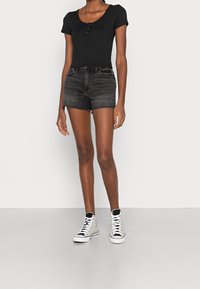 Zwarte geribbelde korte mouw top met drie knopen, gecombineerd met hoge taille donkere spijkerbroek shorts. Schoeisel bestaat uit witte sneakers met zwarte accenten.