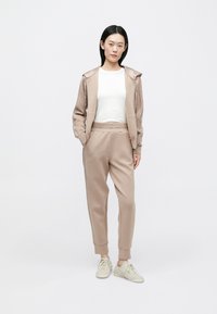 Ανοιχτό καφέ φόρμα με ζακέτα με φερμουάρ και στενά παντελόνια, κατασκευασμένη από απαλό ύφασμα, σε συνδυασμό με ανοιχτά μπεζ πάνινα παπούτσια.