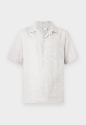 Mann med kort blond hår som har på seg en beige kortermet skjorte med knapper, svarte knelang shorts, hvite sokker og svarte loafers.