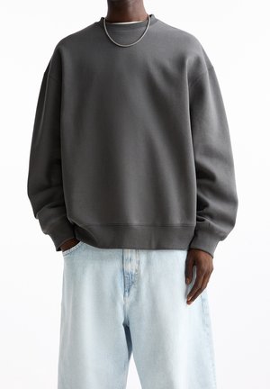 Person trägt dunkelgraues übergroßes Sweatshirt, hellblaue locker sitzende Jeans und eine silberne Kettenhalskette, steht ruhig mit entspannten Händen.