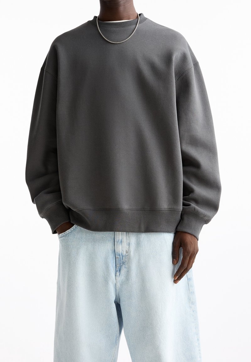 Personne portant un sweat-shirt oversize gris foncé, un jean ample bleu clair et un collier chaîne argenté, debout calmement les mains détendues.