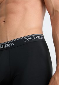 Čierne boxerky vyrobené z hladkej látky, s mäkkým elastickým pásom a bielym logom "Calvin Klein" s dvoma kontrastnými prúžkami.