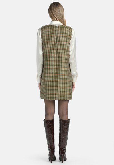 Groene, geruite mouwloze jurk met een hoge halslijn, gecombineerd met een crèmekleurige satijnen blouse en bruine laarzen tot de knie met krokodillenprint.