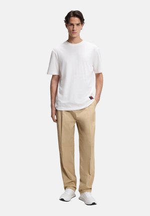 Joven con una camiseta blanca lisa, pantalones beige y zapatillas blancas, de pie contra un fondo claro y liso.