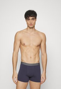 Boxeri din bumbac de culoare bleumarin, cu un talie gri, design ajustat și cusături plate, prezentate pe un model masculin fără cămașă, pe un fundal deschis.