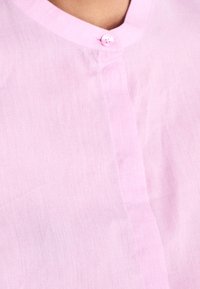 Lekka koszula w jasnoróżowym kolorze, wykonana z cienkiego materiału, z dekoltem w kształcie litery V, pojedynczym zapięciem na guzik i subtelną fakturą. Dopasowany krój, gładka powierzchnia.