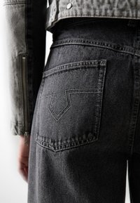 Jeans de denim negro con una vista en primer plano del bolsillo trasero, que presenta un distintivo patrón de costura en forma de V y un acabado texturizado.