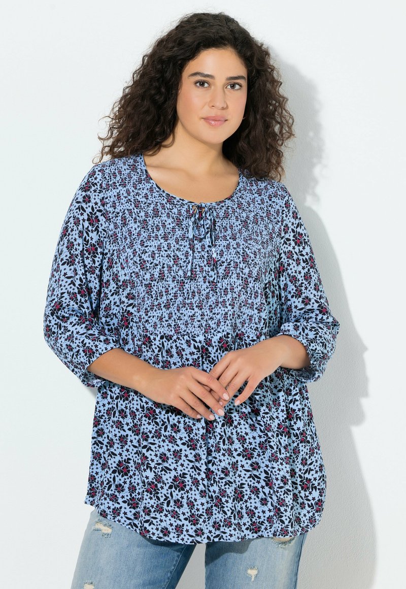 Kwiatowa bluzka w jasnoniebieskim kolorze z czarnymi i czerwonymi wzorami, z okrągłym dekoltem, wiązaniem oraz rękawami do połowy przedramienia.