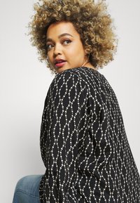 Schwarze Bluse mit weißem gewelltem Punktmuster, runder Ausschnitt und puffärmel. Der Stoff wirkt glatt mit einer leicht glänzenden Textur.