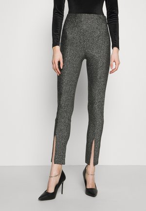 Vrouw draagt sprankelende grijze slim-fit broek met splitjes aan de voorzijde bij de enkels, gecombineerd met zwarte hoge hakken en een zwart topje met lange mouwen.