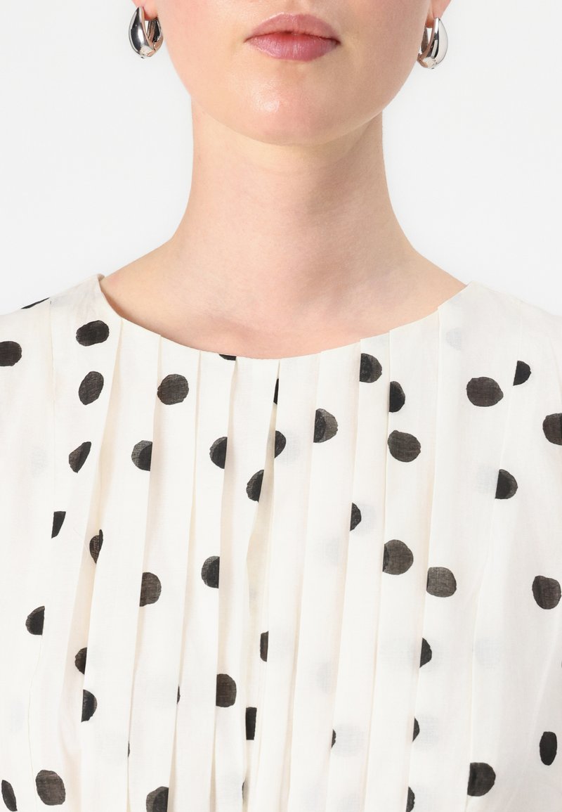 Haut en tissu blanc avec des pois noirs, doté de plis verticaux et d'un col rond. La texture semble légère et lisse.
