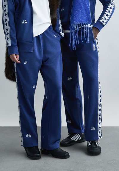 Pantaloni de trening albaștri cu motive de iepuraș alb și dungi laterale cu model de stele. Confecționați din material moale, au croială lejeră și talie elastică.