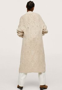 Beżowy sweter na drutach do kolan z wzorem w romby, opuszczonymi ramionami i prążkowanymi mankietami, noszony na białych spodniach i brązowych butach.