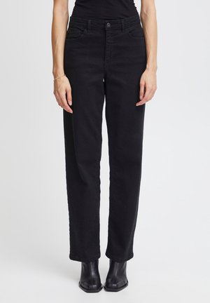 Frau trägt hoch taillierte schwarze Jeans mit geradem Bein und schwarze Stiefeletten, sie steht vor einem schlichten hellen Hintergrund.