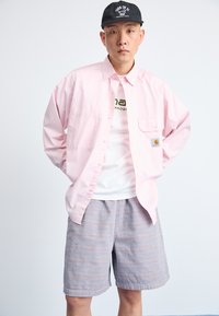 Light pink button-up shirt with pocket, white t-shirt underneath featuring green text, and striped blue shorts. Black cap with text.