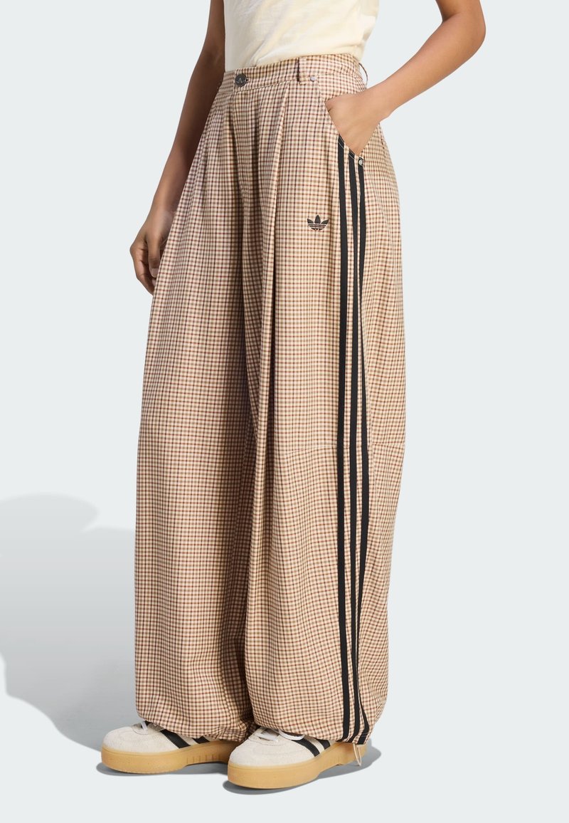 Μπεζ καρό παντελόνια με φαρδύ πόδι, μαύρες πλαϊνές ρίγες και λογότυπο Adidas, φορεμένα με λευκά αθλητικά παπούτσια με μαύρες ρίγες.