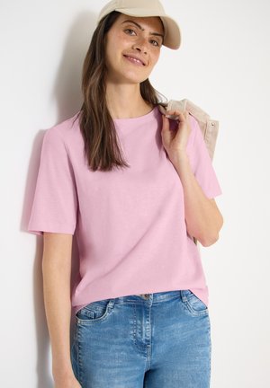 Jeune femme portant un t-shirt rose clair, un jean bleu et une casquette beige, tenant une veste beige sur son épaule sur un fond blanc.