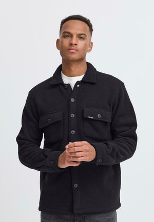 Young man wearing a black button-up fleece jacket with chest pockets, standing against a plain light gray background.