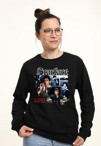 Donna che indossa una felpa nera con diverse scene e testi del film Scarface, con occhiali e capelli raccolti in uno bun.
