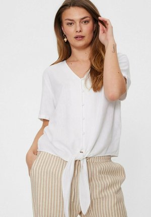 Vrouw met lang haar draagt een wit kort mouwtjes topje met knopen, gestrikt in de taille, en beige broek met witte verticale strepen, staand tegen een witte achtergrond.