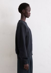 Zwarte oversized sweatshirt met een ronde hals, ribgebreide manchetten en subtiele logodetails. Gladde textuur, gedragen met een lange donkere rok.