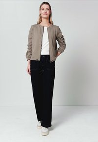 Frau steht vor schlichtem hellem Hintergrund und trägt eine taupefarbene, gesteppte Jacke, ein weißes Hemd, schwarze Hosen mit weitem Bein und weiße Sneakers.