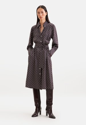 Femme portant une robe noire à manches longues à pois blancs, taille nouée, et des bottes noires à talons montant jusqu'aux genoux, debout les mains dans les poches.