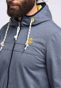Grå zip-up hoodie med en mjuk textur, med gula accenter, trädragkedjor och dekorativ sömnad på bröstet.
