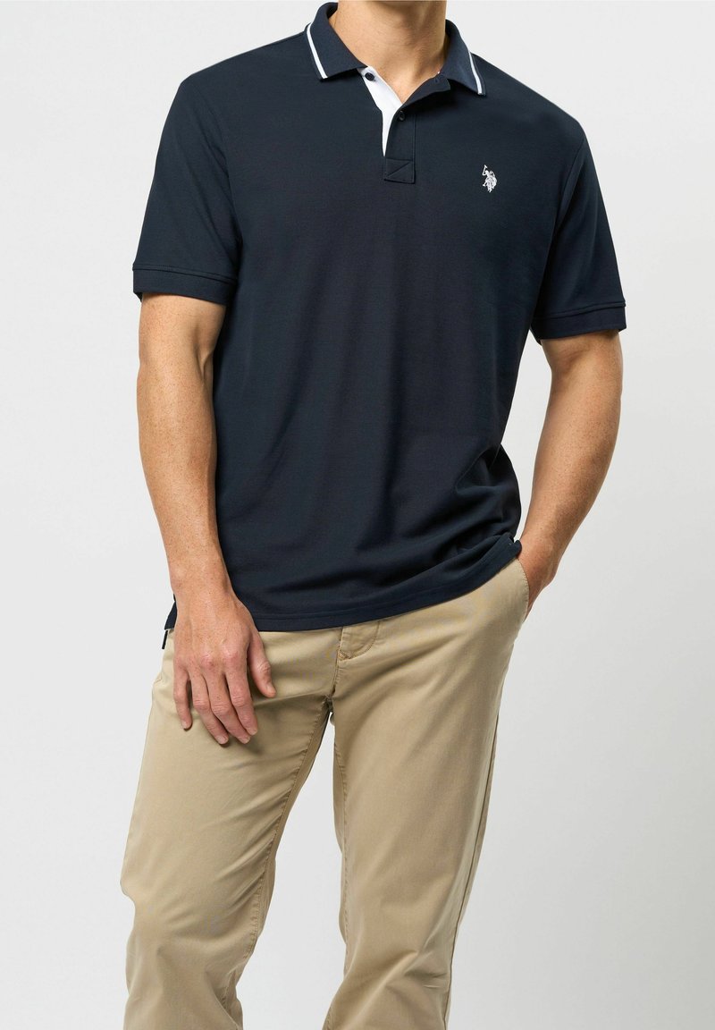 Homme portant un polo bleu marine avec un col blanc et un pantalon beige, debout avec une main dans la poche devant un fond uni.