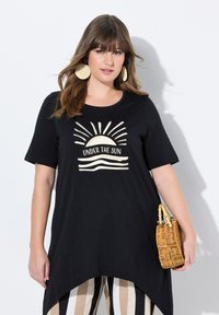 Camiseta negra con un gráfico frontal que presenta un sol y olas, de mangas cortas y con un dobladillo asimétrico. Combinada con pantalones a rayas y un bolso de mimbre.
