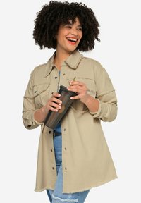 Giacca camicia in cotone beige con bordi sfrangiati, quattro tasche, bottoni frontali e leggero curvatura all'orlo. Il modello tiene una tazza nera.