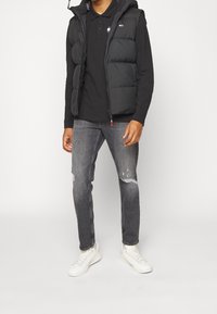 Svart pufferväst över en svart långärmad tröja, kombinerad med slitna gråjeans och vita sneakers. Funktioner inkluderar dragkedjor och logodetaljer.