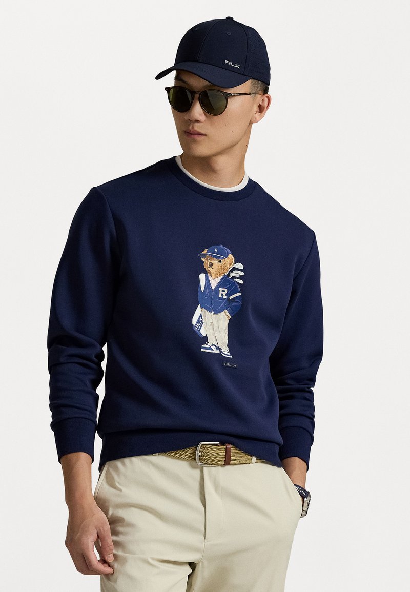 Pull en marine avec une illustration d'ours en tenue de sport, col rond, poignets et ourlet côtelés. Associé à un pantalon kaki et une casquette.