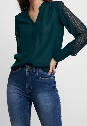 Femme portant un chemisier bleu sarcelle foncé boutonné avec des manches en dentelle et un jean taille haute bleu, debout avec une main sur la hanche.