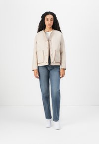 Beige vadderad jacka med rund halsringning, framknäppning, två sidofickor och kontrasterande kant. Bärs över en vit skjorta och blå jeans.