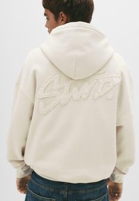 Cremefarbener Hoodie mit einem großen, strukturierten Logo, das auf den Rücken gestickt ist. Er verfügt über eine Kapuze mit Kordelzug und gerippte Bündchen, kombiniert mit blauen Jeans.