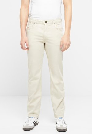 Personne portant un pantalon droit écru, des baskets blanches avec des bandes noires et un haut blanc sans manches, debout devant un fond uni.