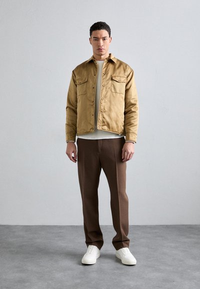 Tan bomber jacket with two chest pockets, a beige sweater underneath, brown trousers, and white sneakers. Smooth texture, fitted design.