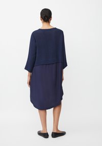 Frau mit dunklen Haaren im Dutt, die ein navyblaues, geschichtetes Kleid und schwarze Ballerinas trägt, steht vor einem schlichten weißen Hintergrund, Rückansicht.