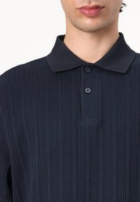 Navy ribbed polo shirt with a pointed collar, three black buttons, and textured fabric, focusing on vertical lines and smooth texture.