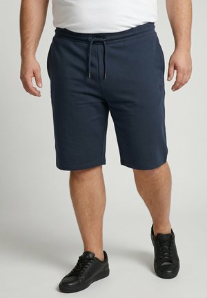 Uomo che indossa pantaloncini blu navy con coulisse, t-shirt bianca e sneakers nere in piedi su uno sfondo semplice.