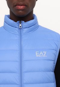 Svetle modrá péřová vesta s vysokým límcem, zipovým zapínáním a logem EA7 Emporio Armani. Disponuje prošitými panely a hladkým povrchem.